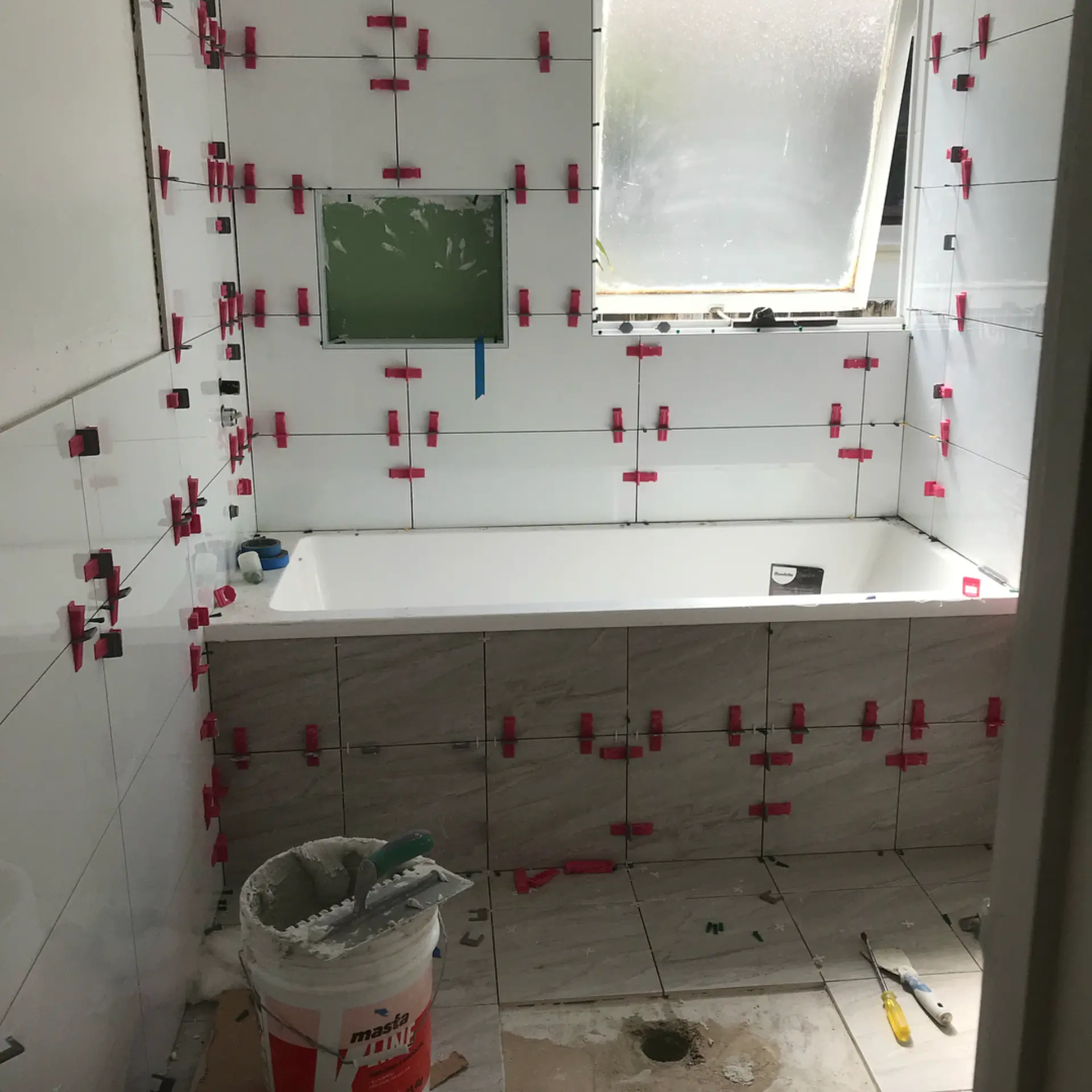 Bathroom tiling in progress with levelling clips on white wall tiles and timber-look bath surround